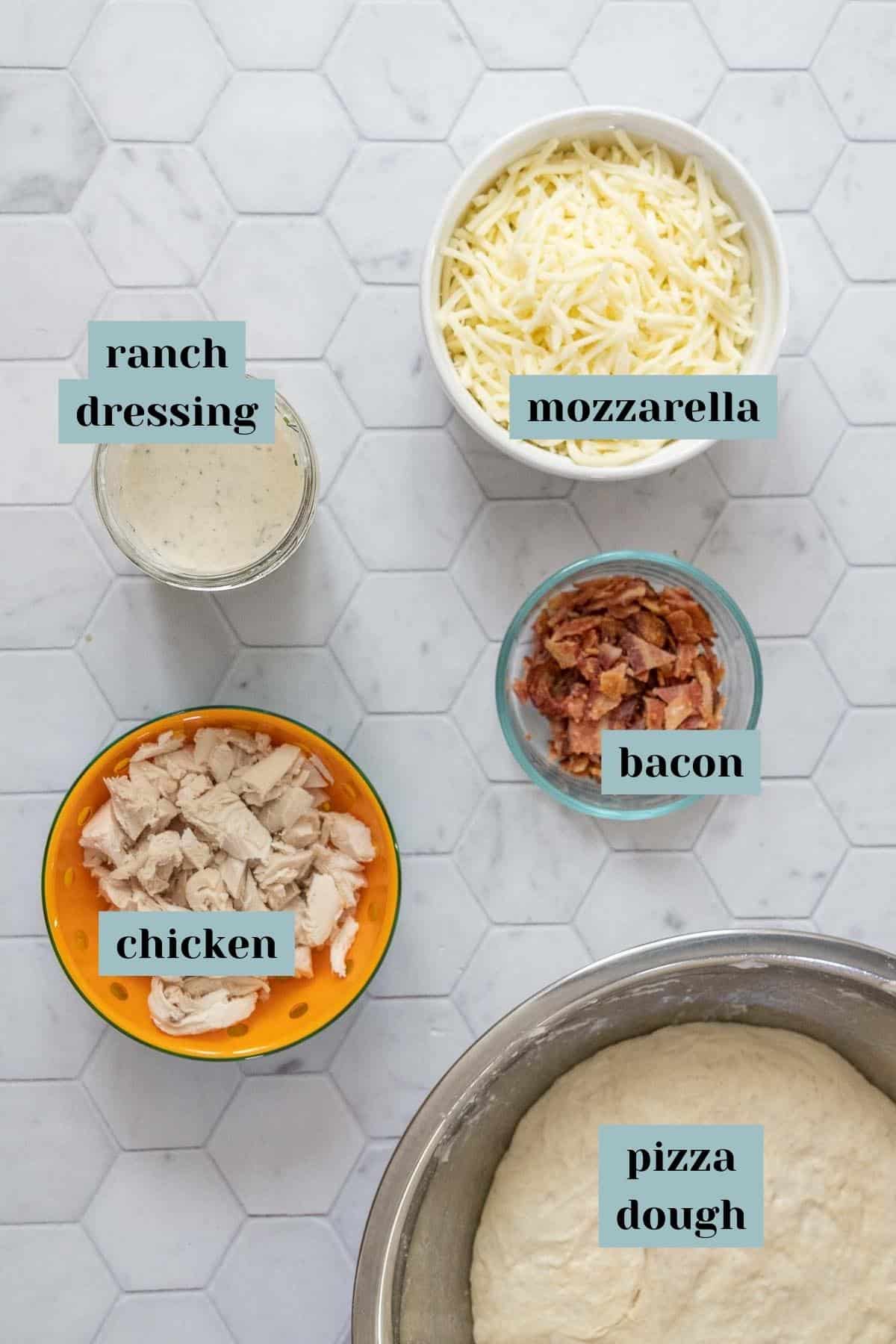 Ingredients for chicken bacon ranch pizza on tile surface with labels.