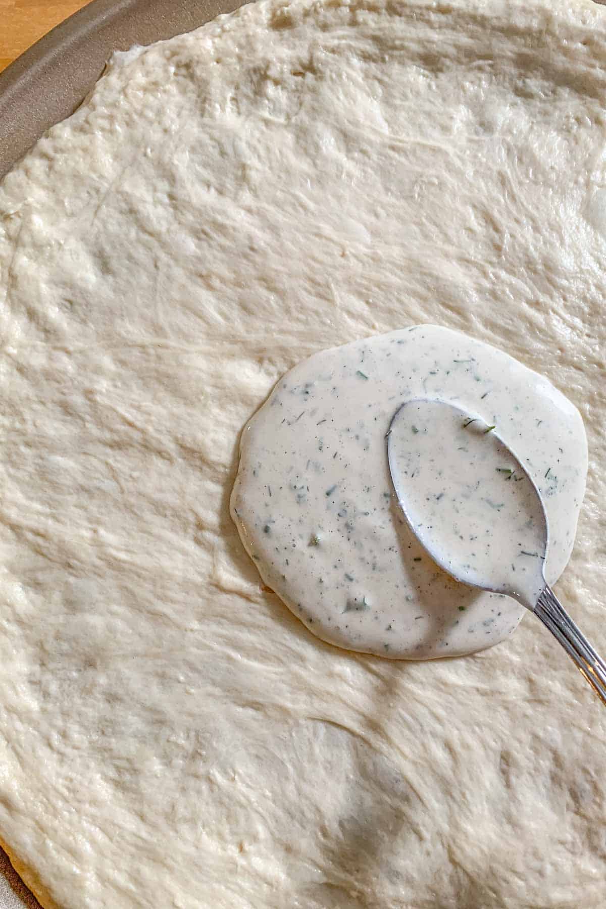 Unbaked pizza crust with ranch dressing being spread on with a spoon.