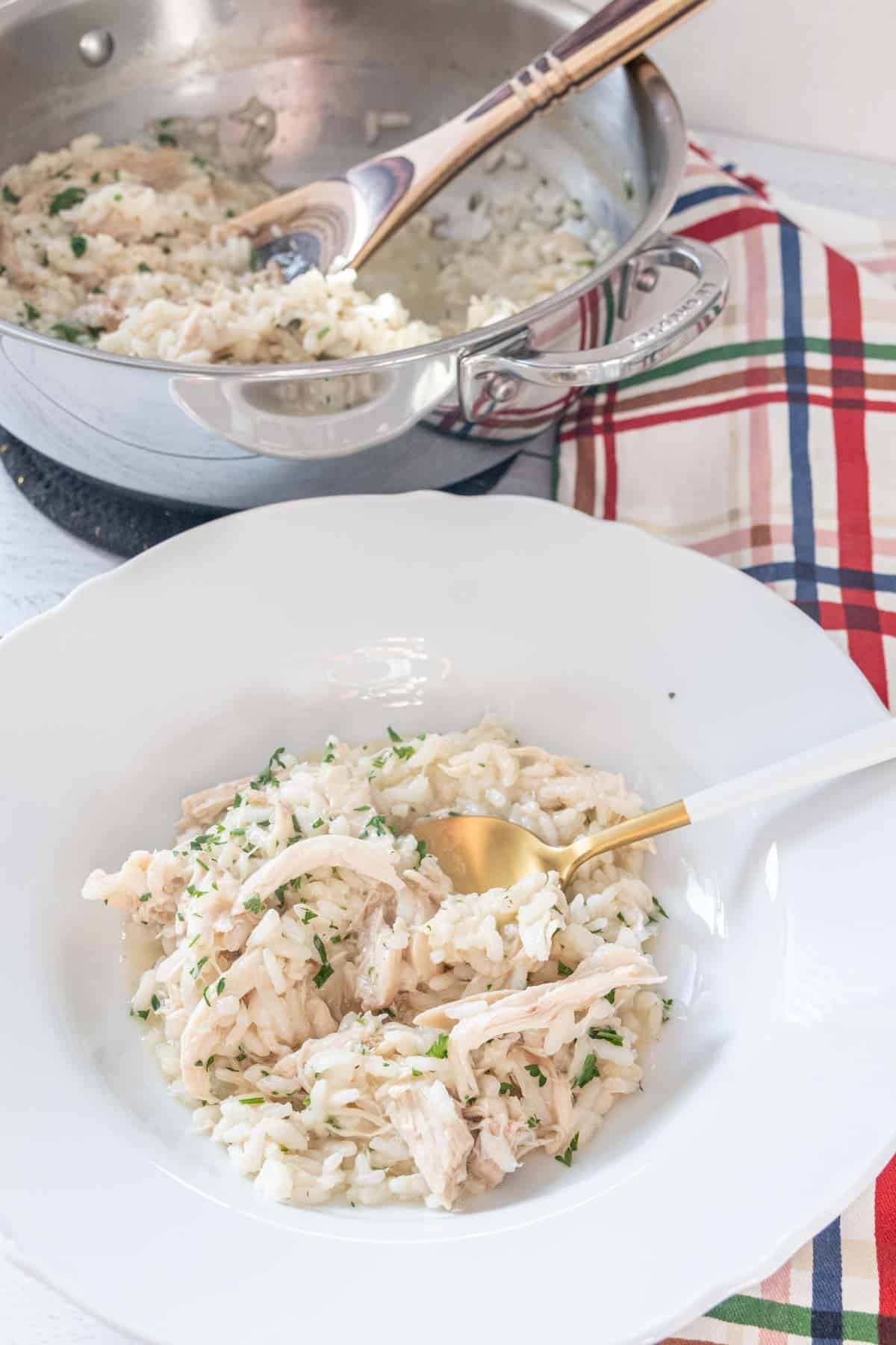 White bowl of chicken risotto with pan of risotto behind.