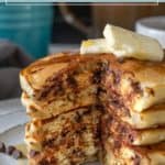 A stack of fluffy chocolate chip pancakes with a pat of butter on top sits on a plate, a bite taken out and extra chocolate chips scattered nearby.