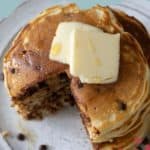 A stack of fluffy chocolate chip pancakes topped with two pats of butter and a drizzle of syrup, with one wedge cut out, sits invitingly on a white plate.