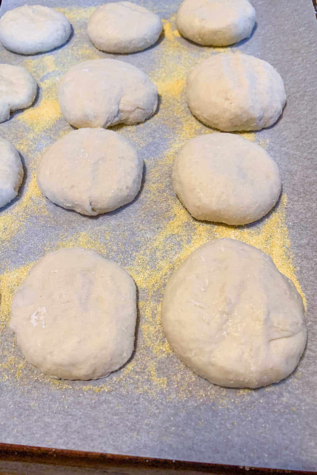 English muffin dough balls resting on a baking sheet covered in cornmeal.