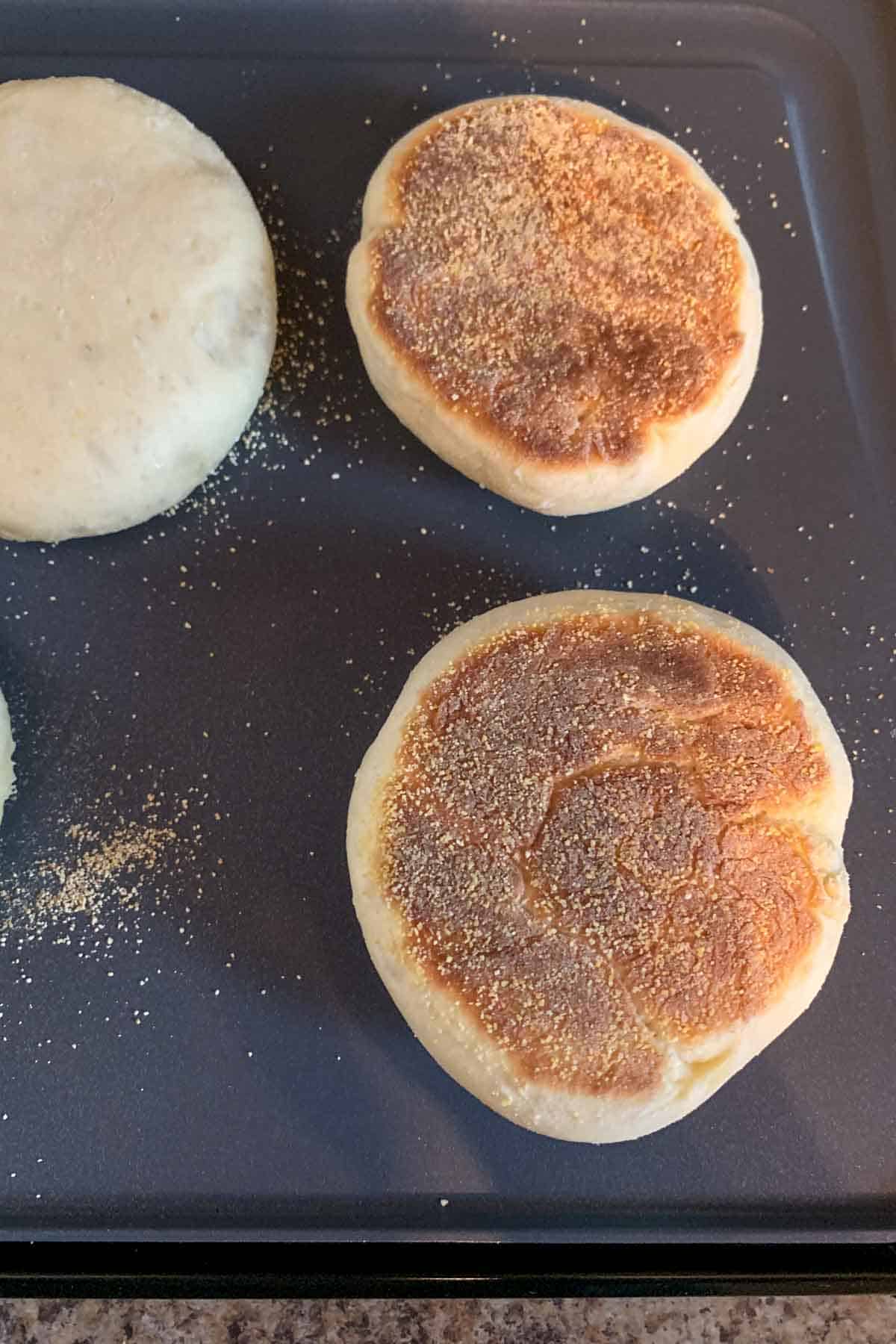 Flipped English muffin on a griddle showing one side baked.