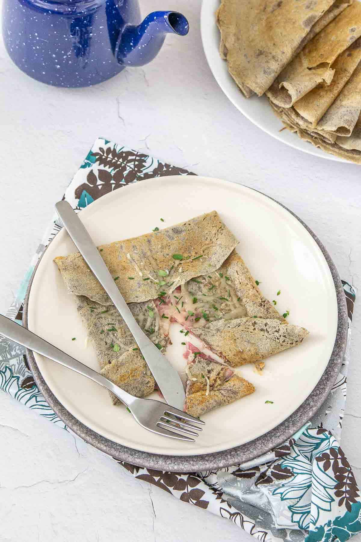 Gluten free crepe with ham and cheese on a plate, cut open with knife and fork.