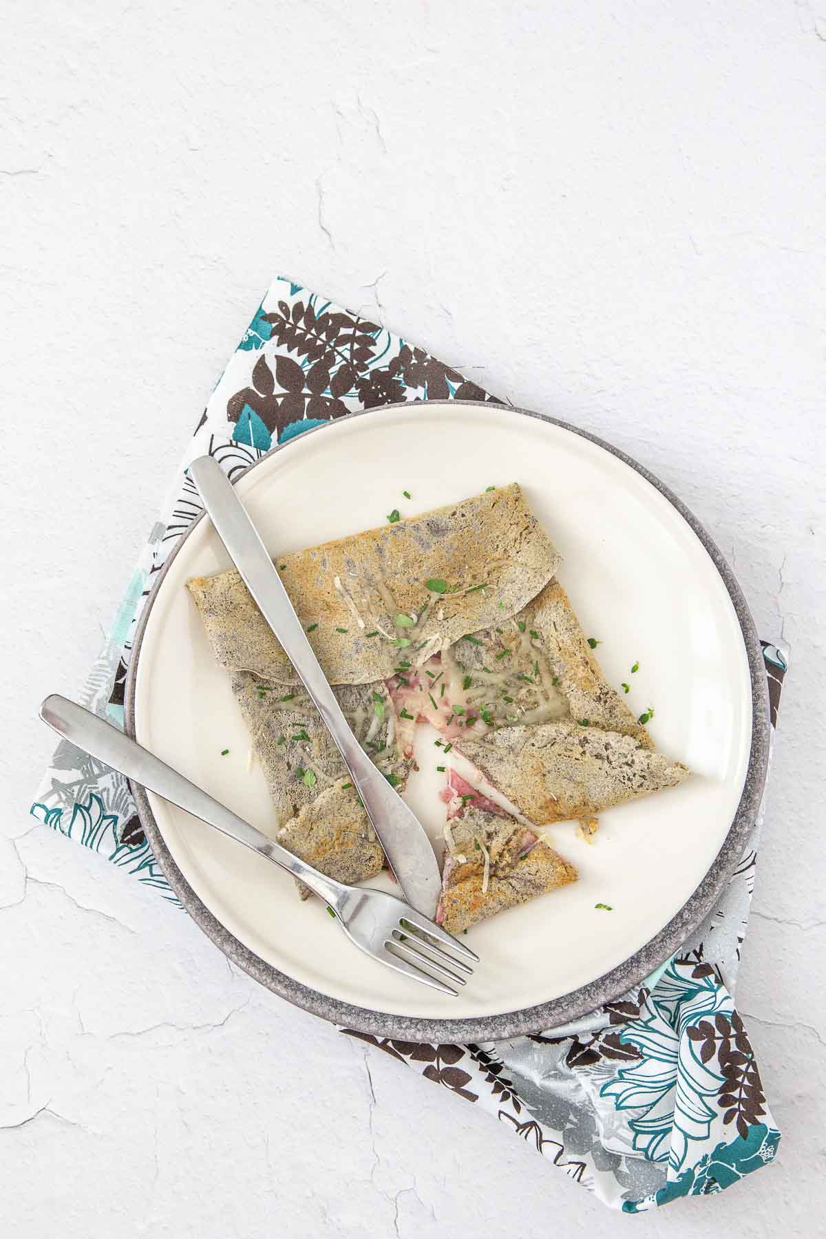 Overhead photo of a gluten free crepe filled with ham and cheese on two stacked plates with a floral napkin.