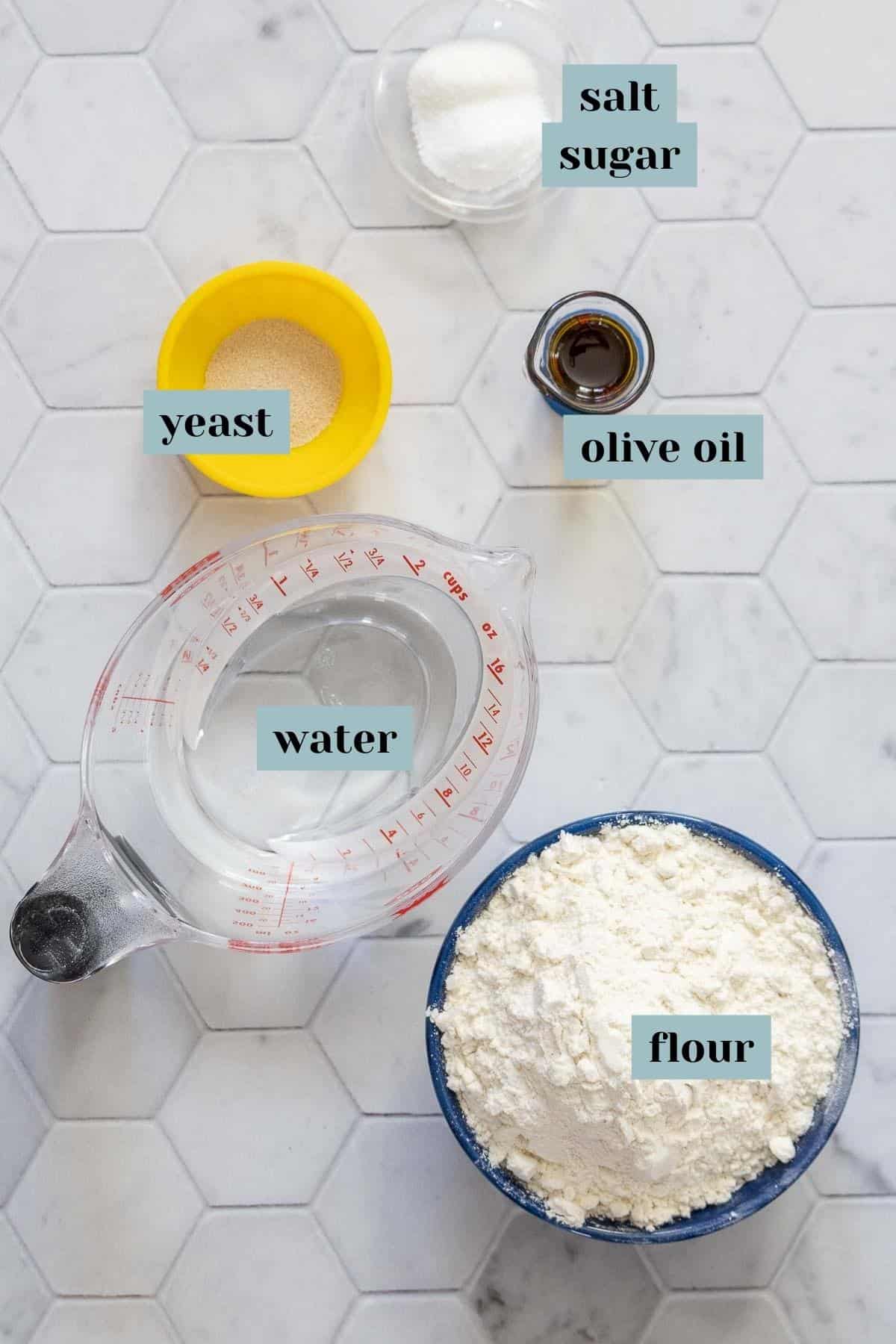Ingredients for homemade pizza dough on a tile surface with labels.