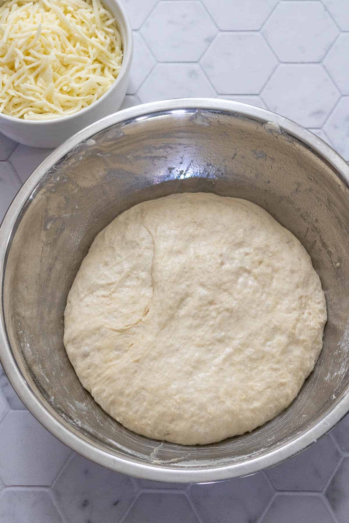 Metal bowl of risen pizza bowl with a bowl of cheese next to it.