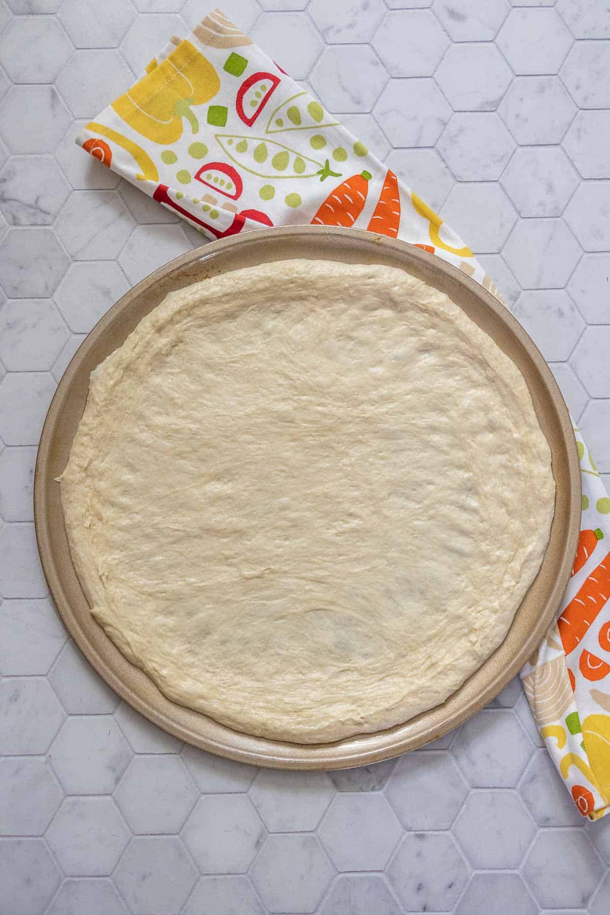 Unbaked pizza dough on a pizza pan with a printed kitchen towel.