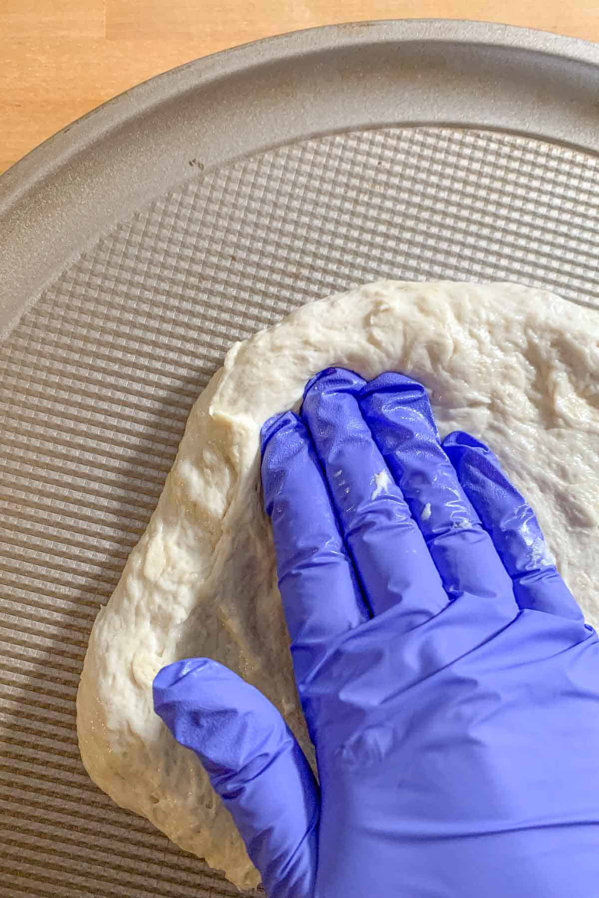 Gloved hand pressing pizza dough onto pan.