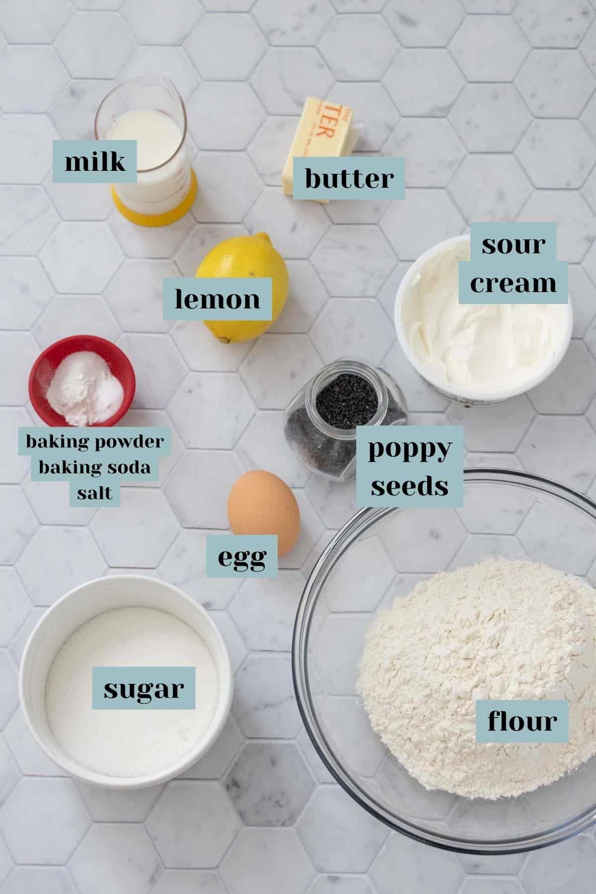 Ingredients for lemon poppy seed bread on a tile surface with labels.