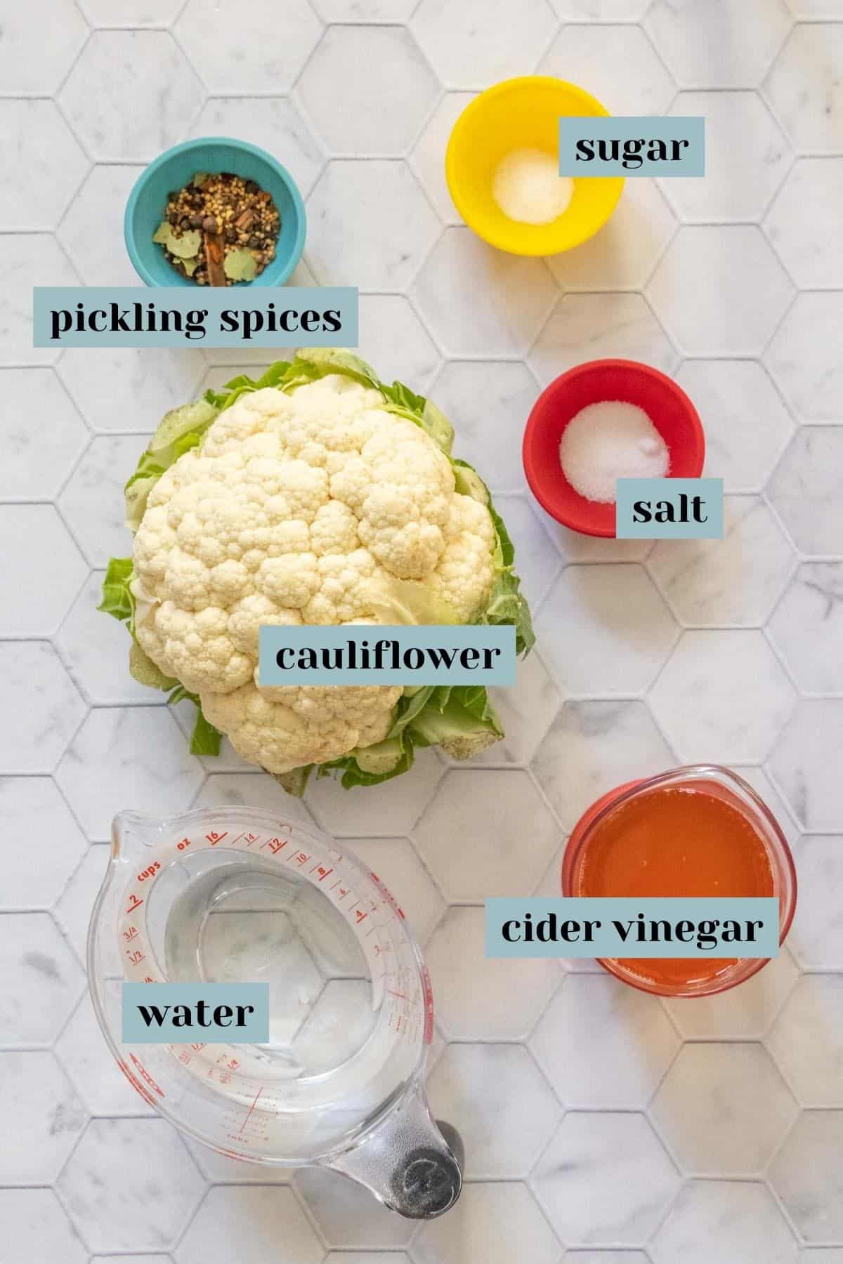 Ingredients for pickled cauliflower on a tile surface with labels.