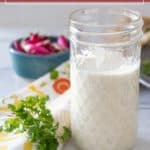 A glass jar filled with creamy ranch dressing sits on a table, surrounded by fresh parsley, a patterned cloth, and a bowl of chopped radishes in the background.