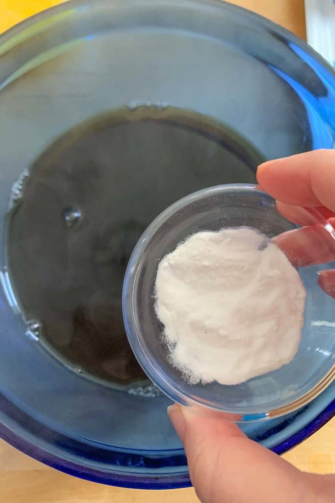 Hand holding a small bowl of baking soda over a blue mixing bowl containing molasses and water.
