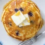 Overhead of plate of blueberry pancakes with butter and syrup on top with a fork beside.