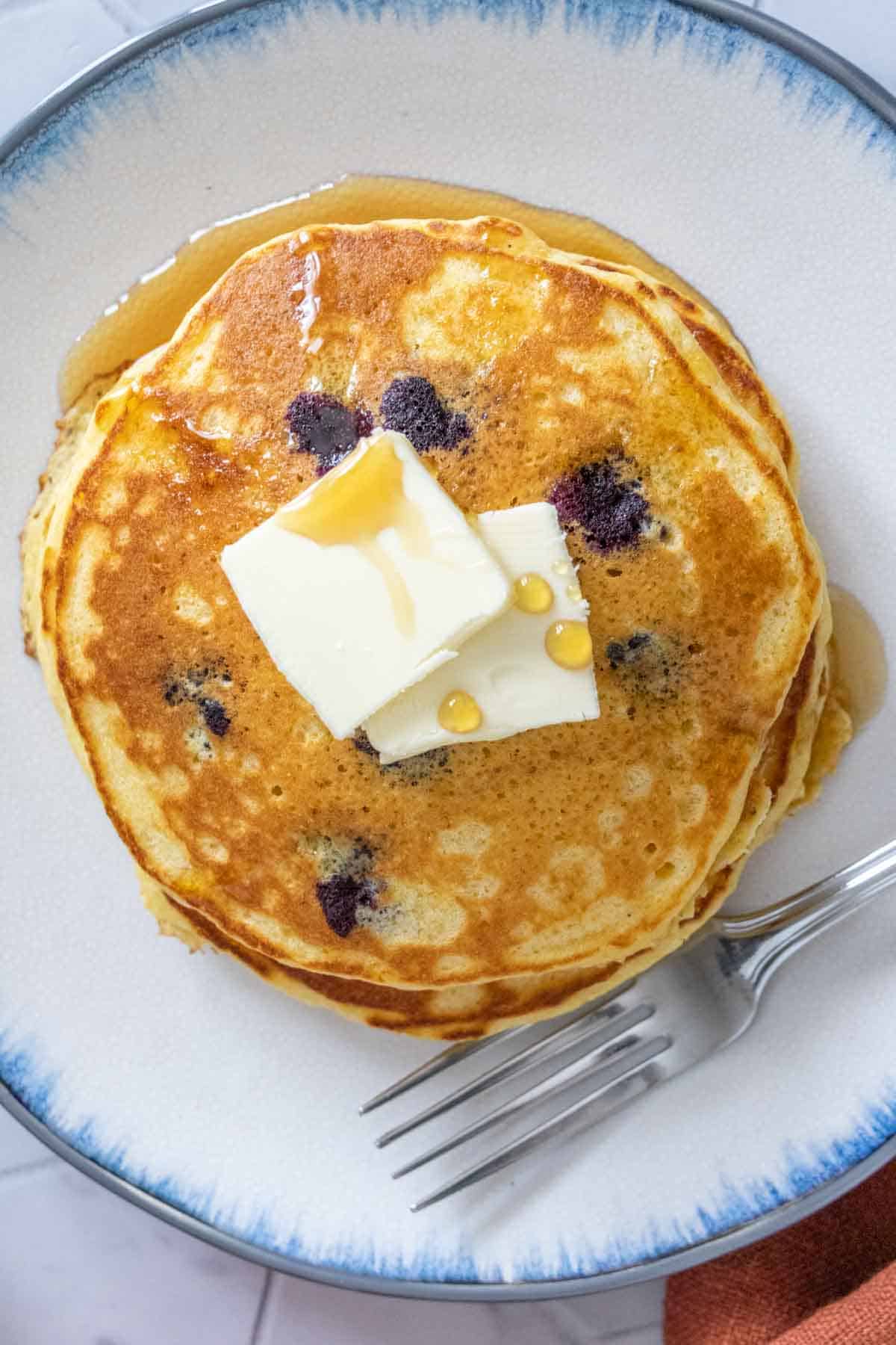 Overhead of plate of blueberry pancakes with butter and syrup on top with a fork beside.