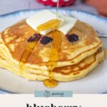 A stack of fluffy blueberry pancakes topped with butter and syrup being poured from a small blue pitcher.