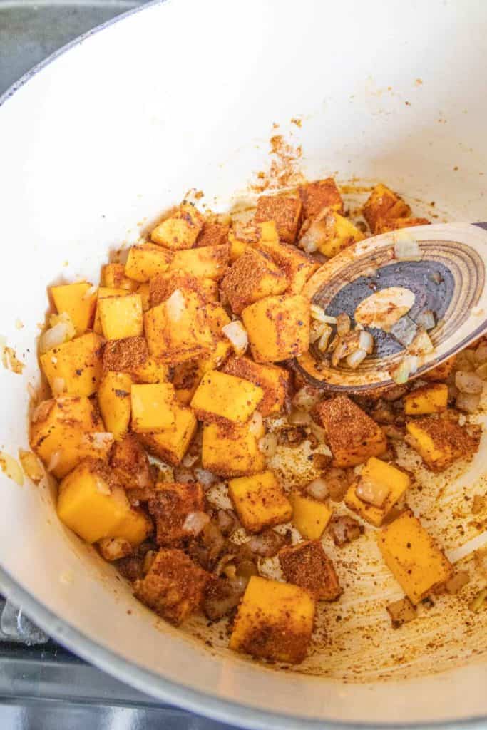 Butternut squash, onion, and spices in dutch oven being stirred with wooden spoon.