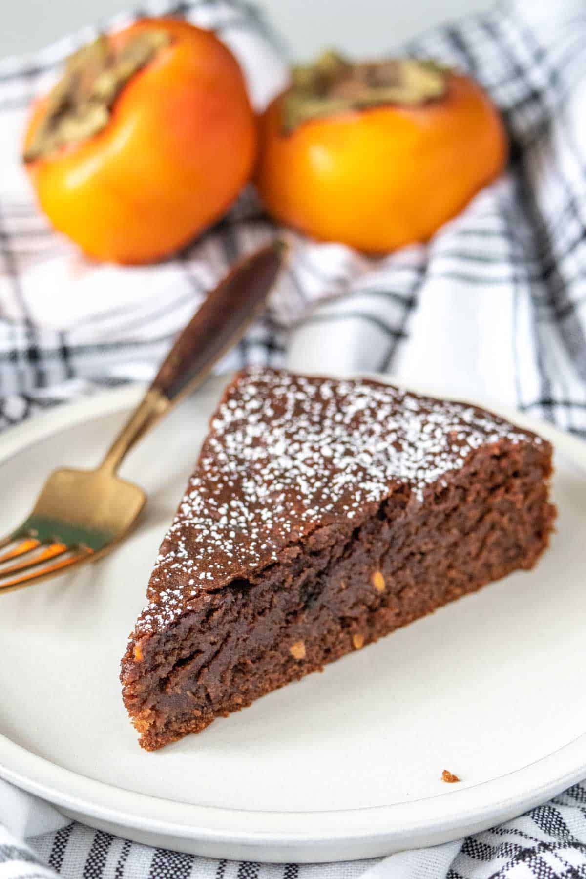 Slice of chocolate persimmon cake on a plate with a fork and persimmons behind.