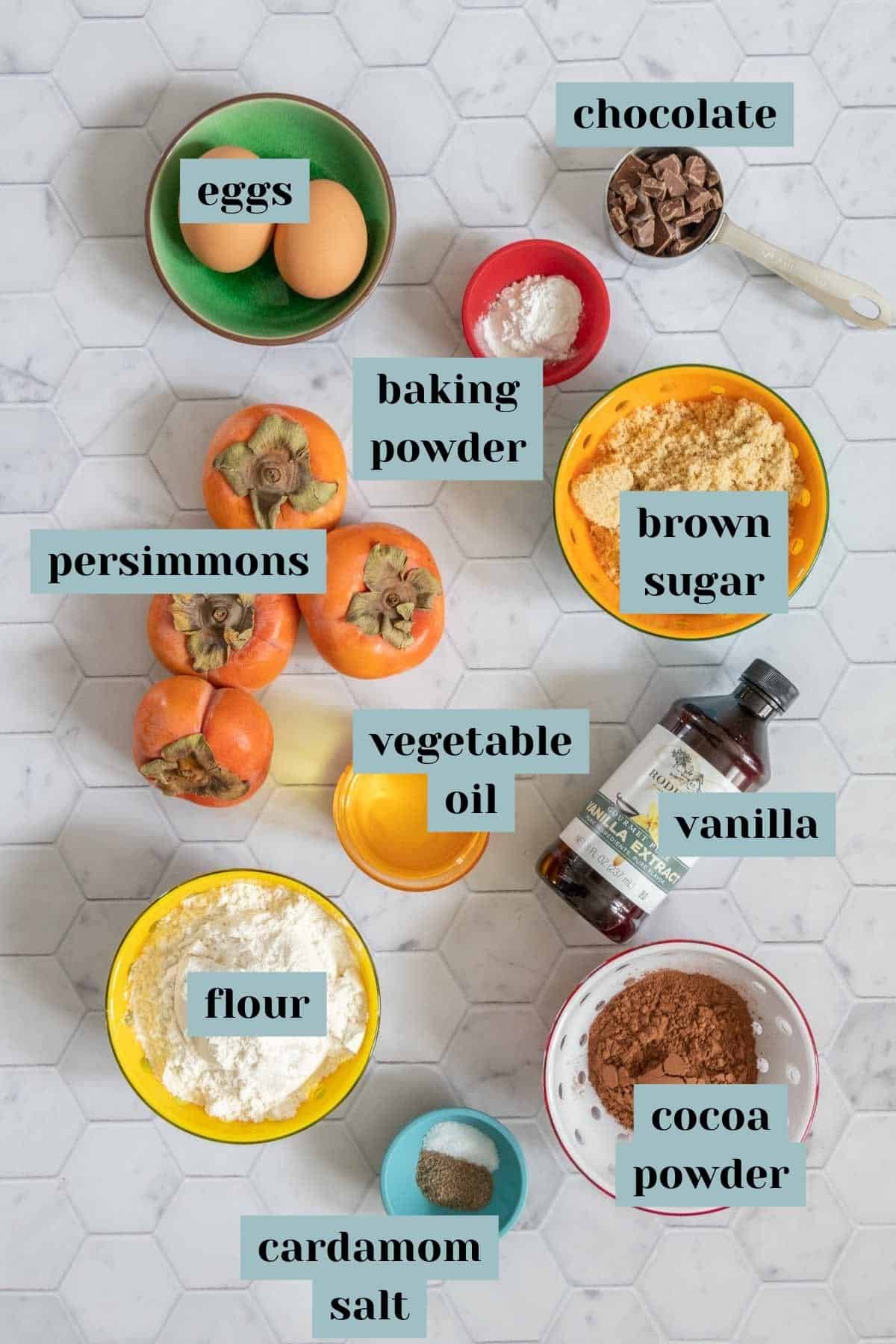 Ingredients for chocolate persimmon cake on a tile surface with labels.