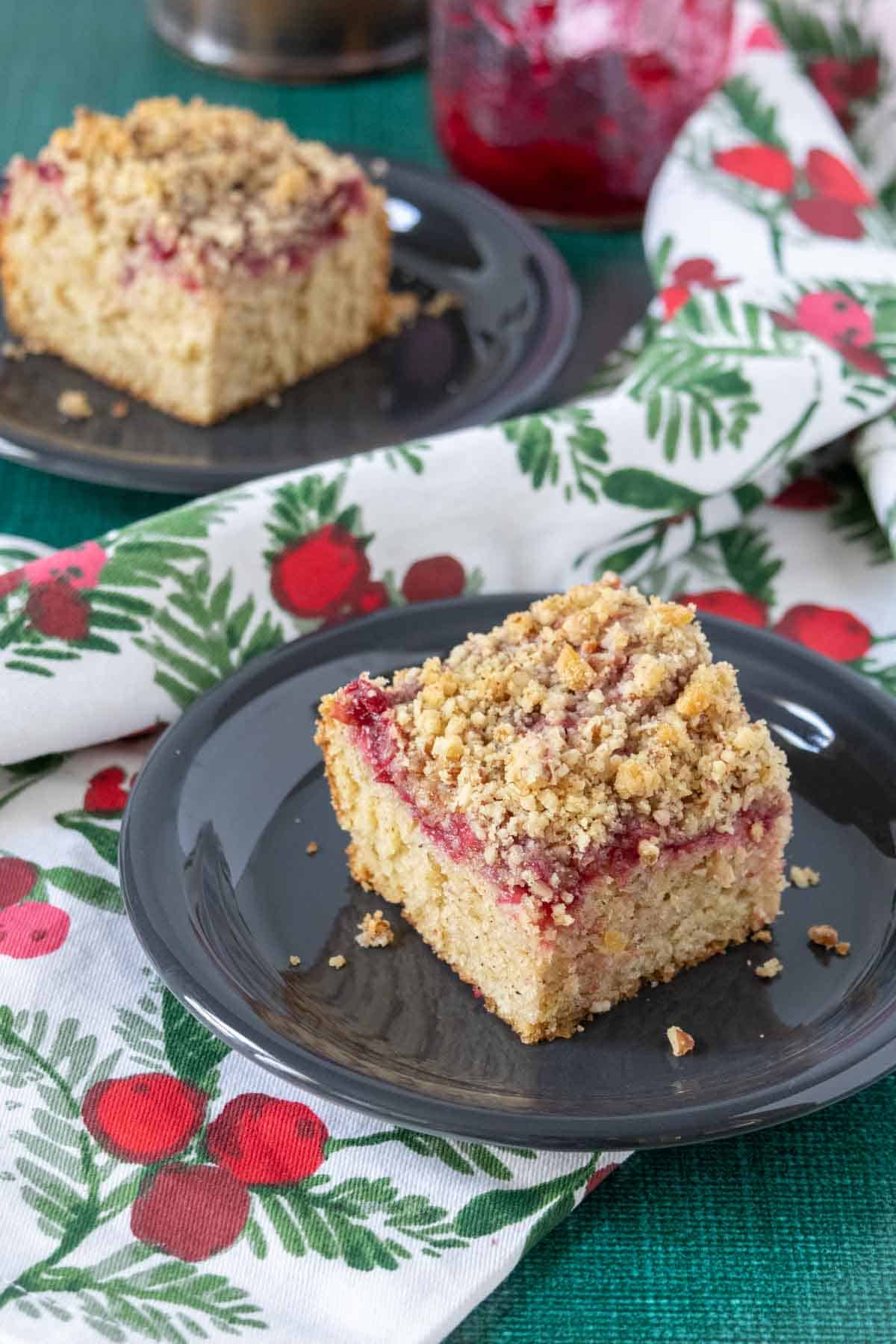 Slice of cranberry coffee cake on a black plate with a cranberry print napkin.