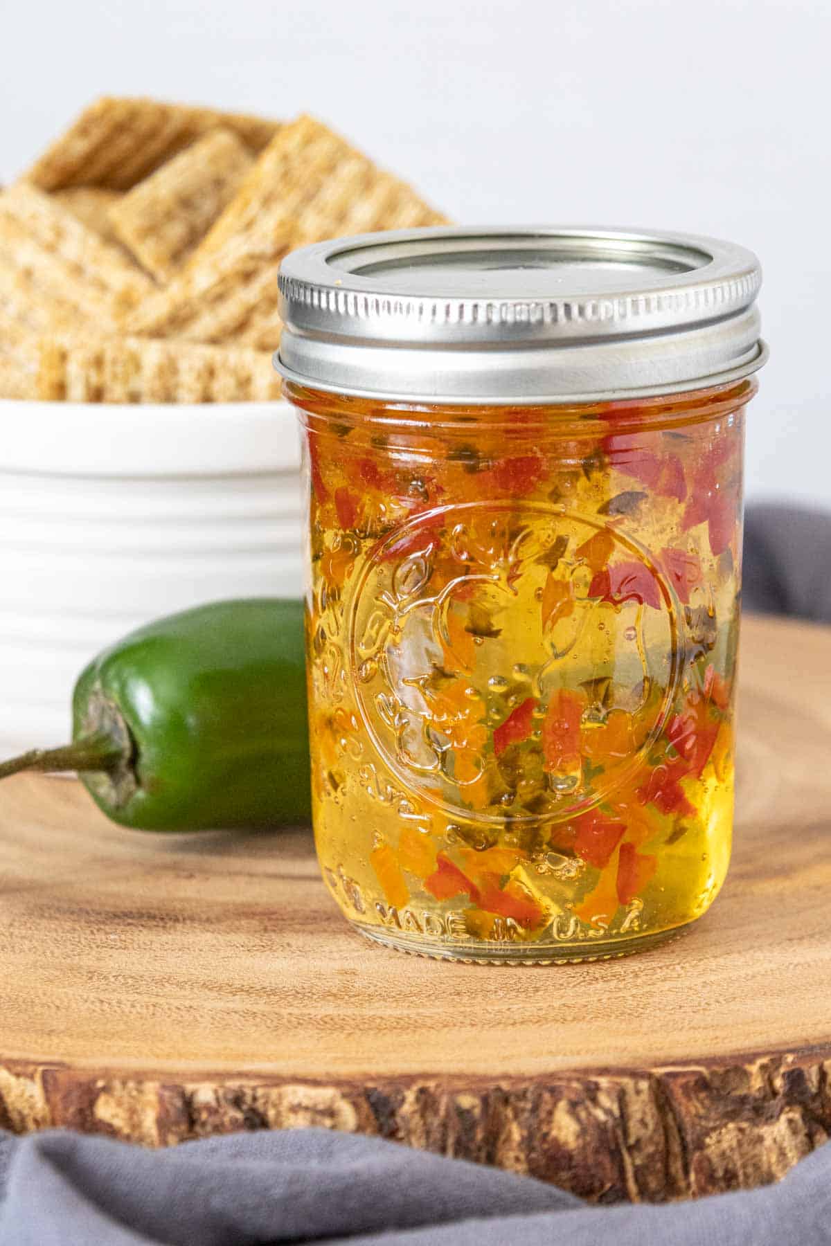 Jar of jalapeño jelly on a wood tray with a jalapeño and crackers behind.