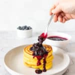 Blueberry sauce being spooned over a stack of cornmeal pancakes on a gray plate.