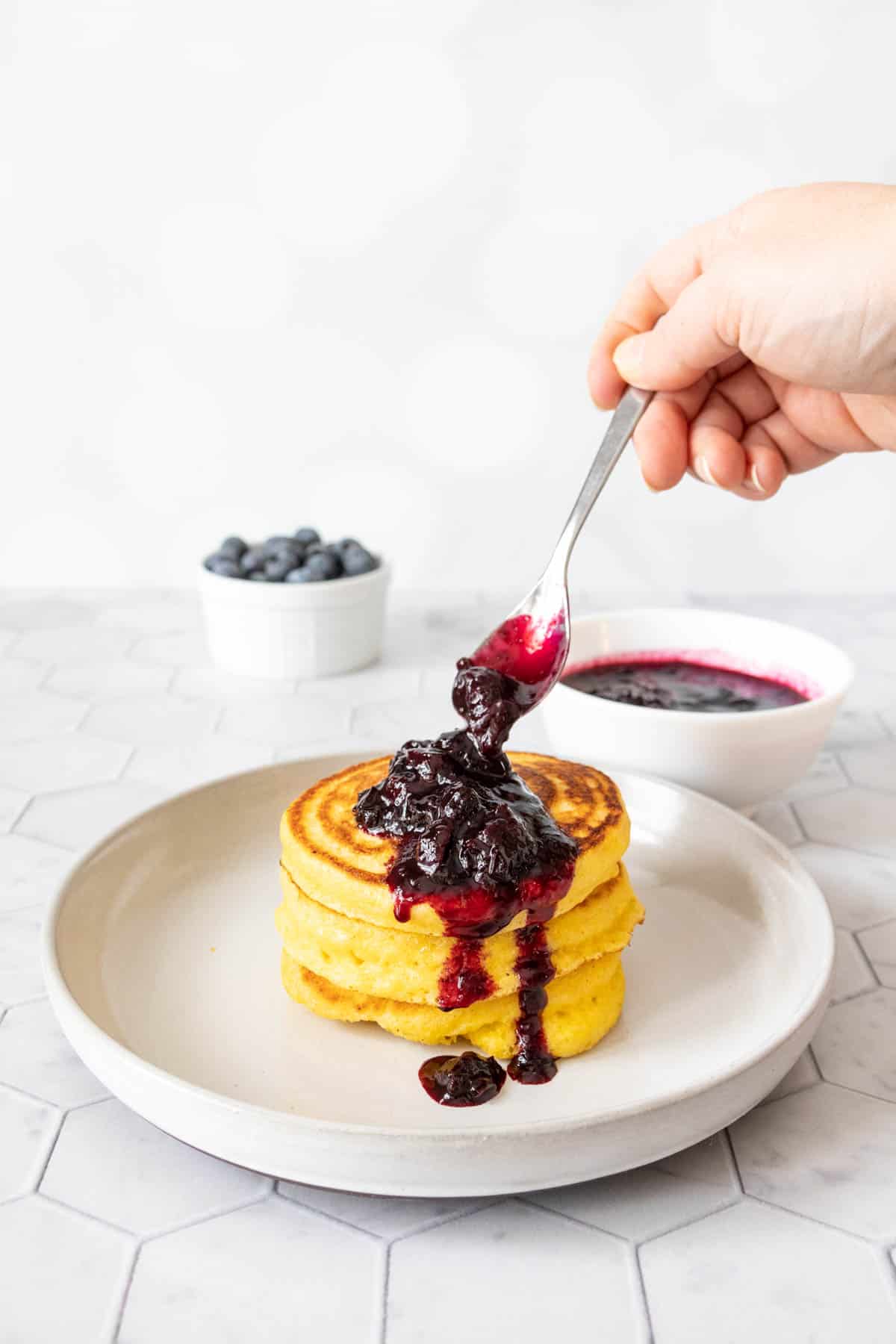 Blueberry sauce being spooned over a stack of cornmeal pancakes on a gray plate.