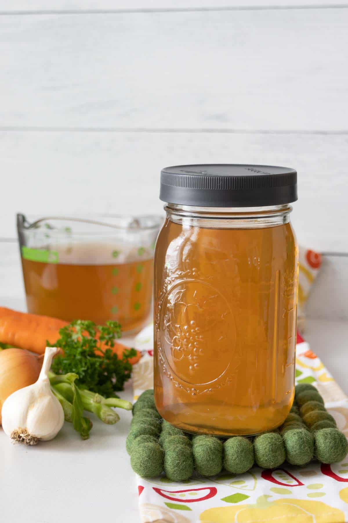 Jar of homemade vegetable stock on a green felt trivet with measuring cup of broth behind.