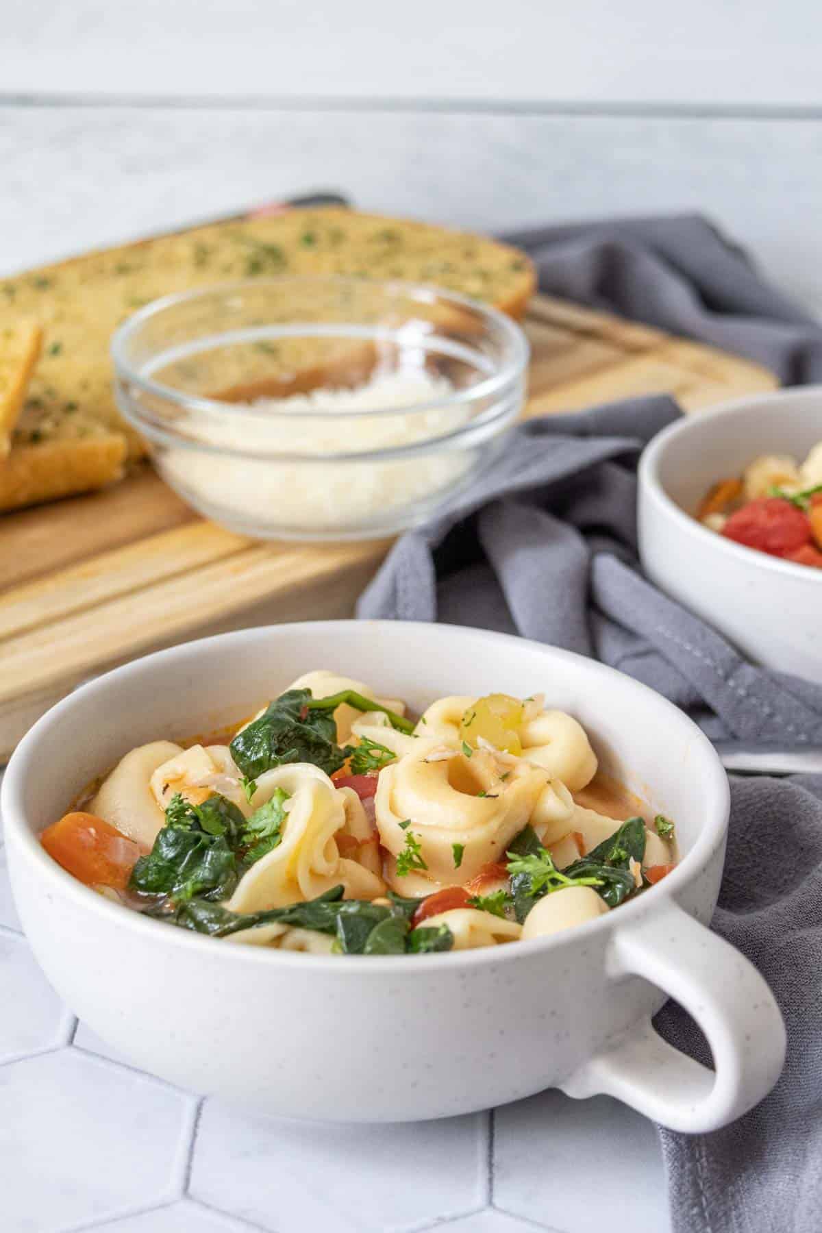 Low mug filled with tortellini soup and garlic bread in the background.