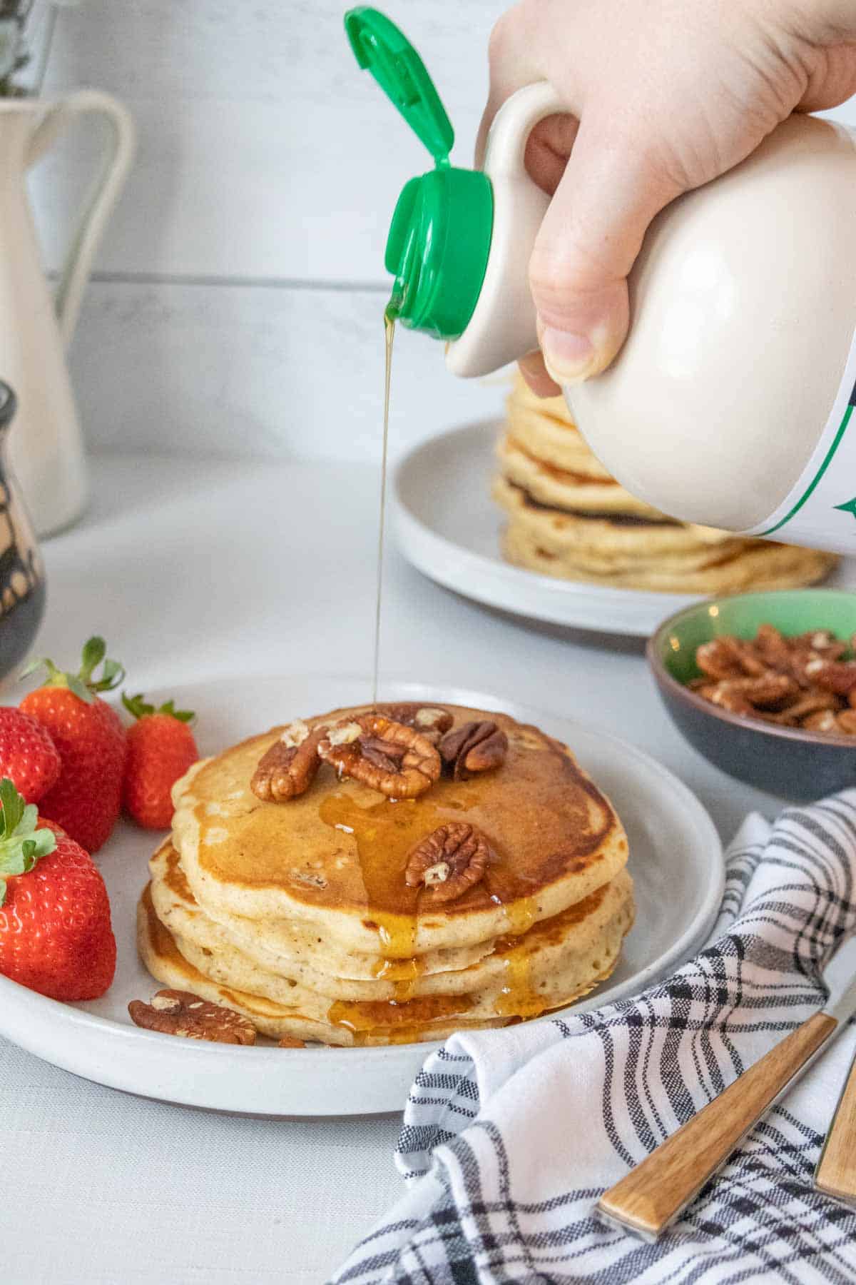 Caucasian hand pouring syrup from a bottle onto a stack of pecan pancakes.