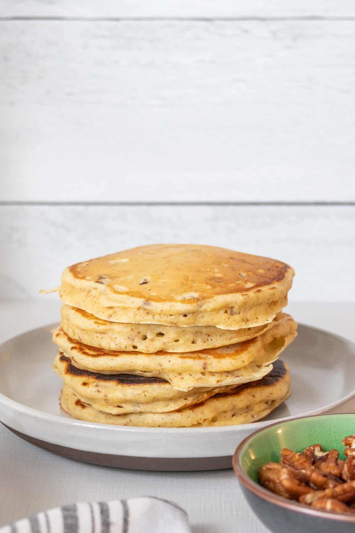 Stack of pecan pancakes on a gray plate with pecan pieces in a green bowl beside.