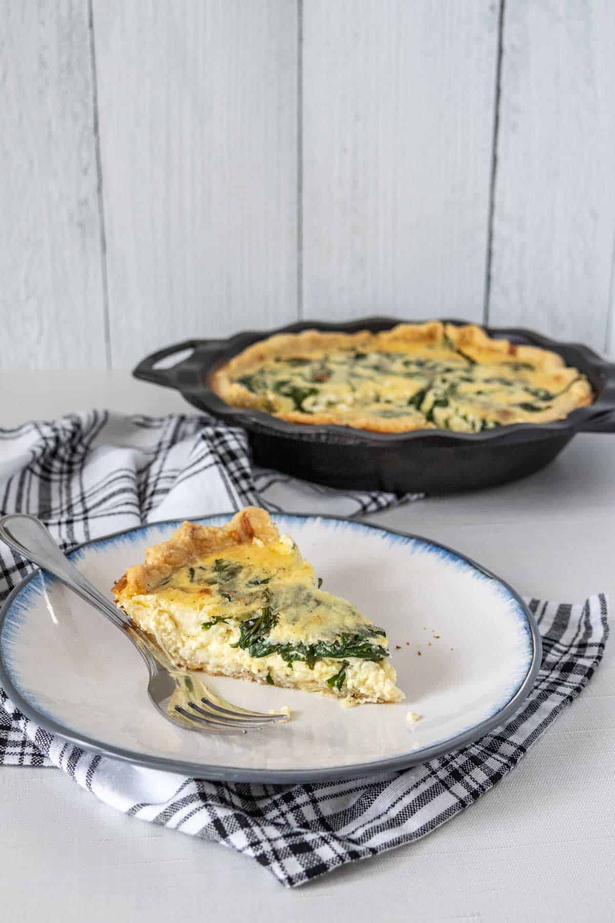 Slice of spinach quiche on a plate with rest of quiche behind.