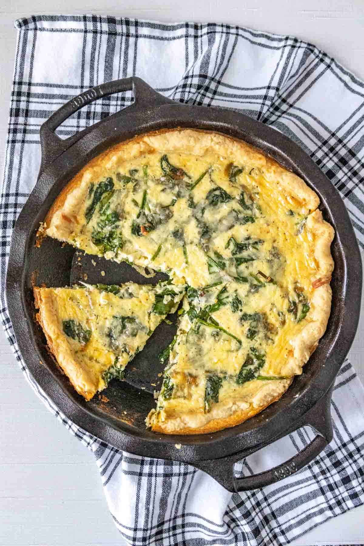 Spinach quiche in a cast iron pie plate with a black and white kitchen towel.