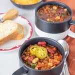 Mugs of three bean chili with spoons, bread, and shredded cheese beside.