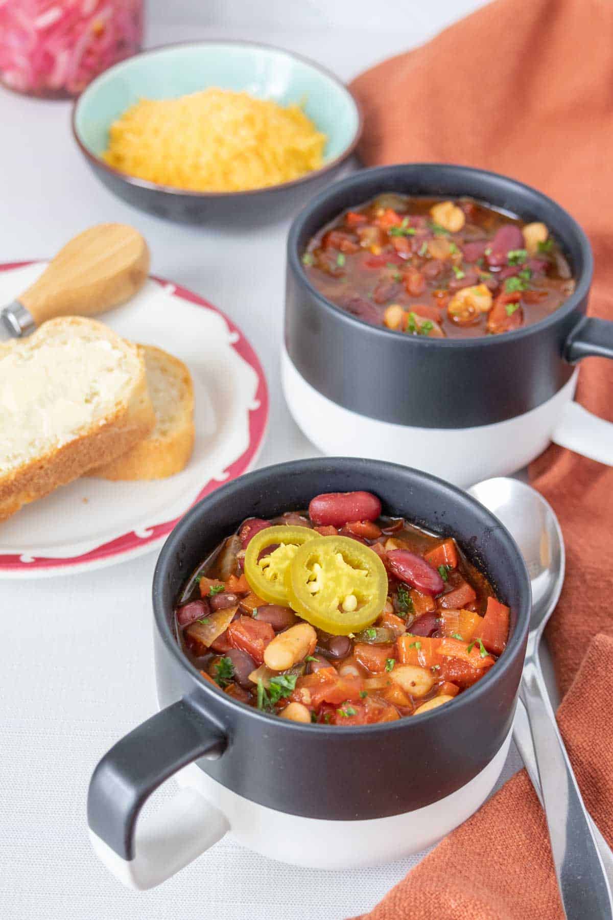 Mugs of three bean chili with spoons, bread, and shredded cheese beside.