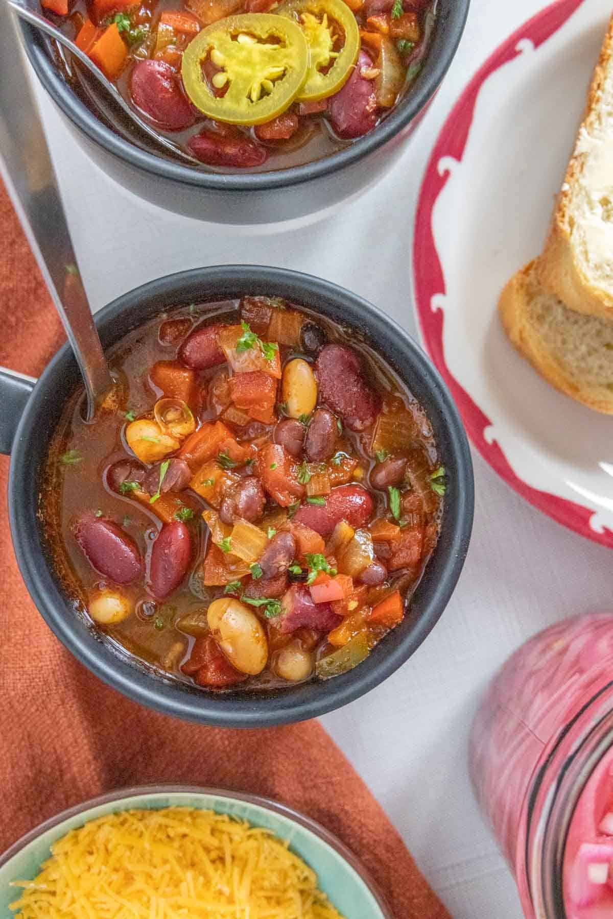 Overhead of mugs of three bean chili with bread, cheese, and pickled onions off the sides.