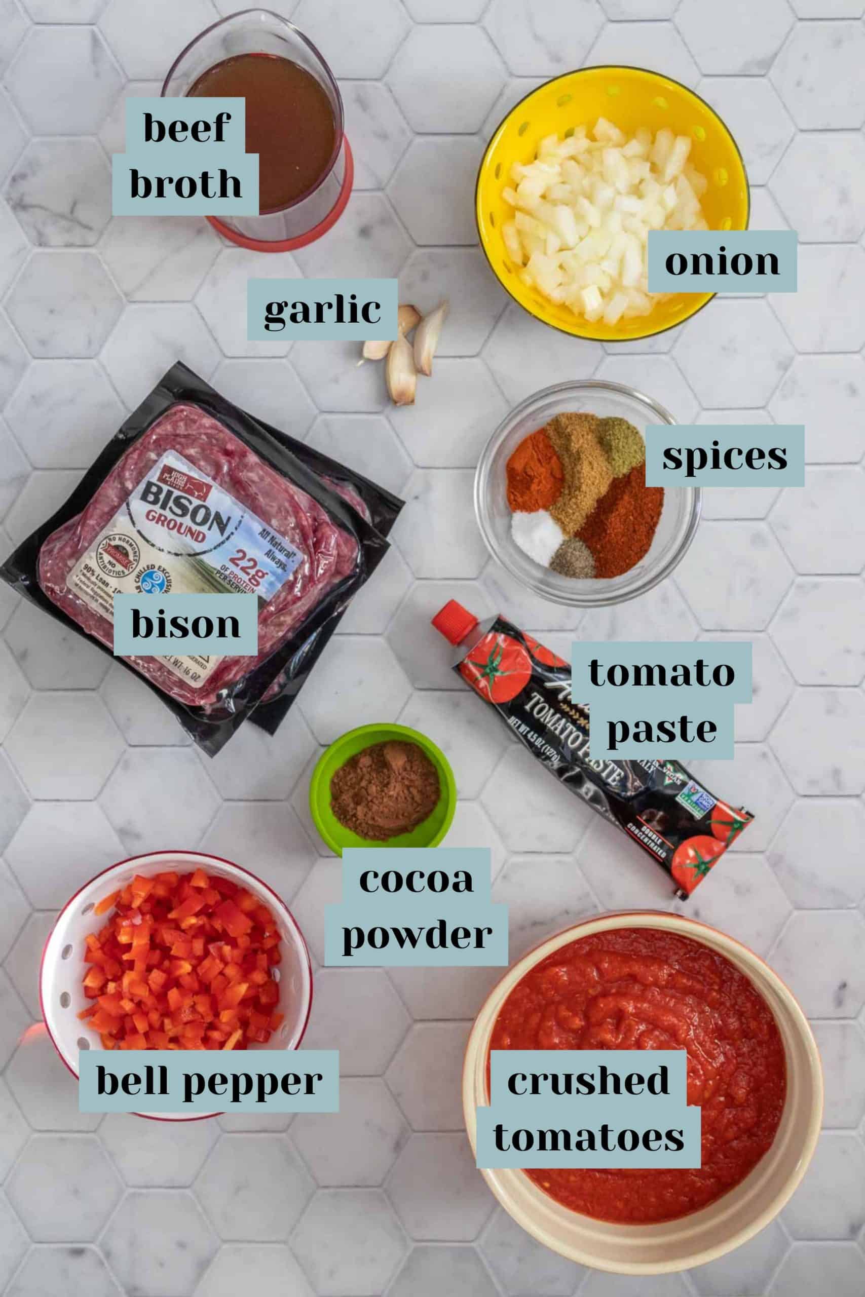 Ingredients for bison chili on a tile surface with labels.