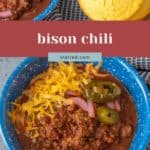 A blue bowl of hearty bison chili topped with shredded cheese, pickled onions, and jalapeños sits next to a muffin on a black and white cloth.