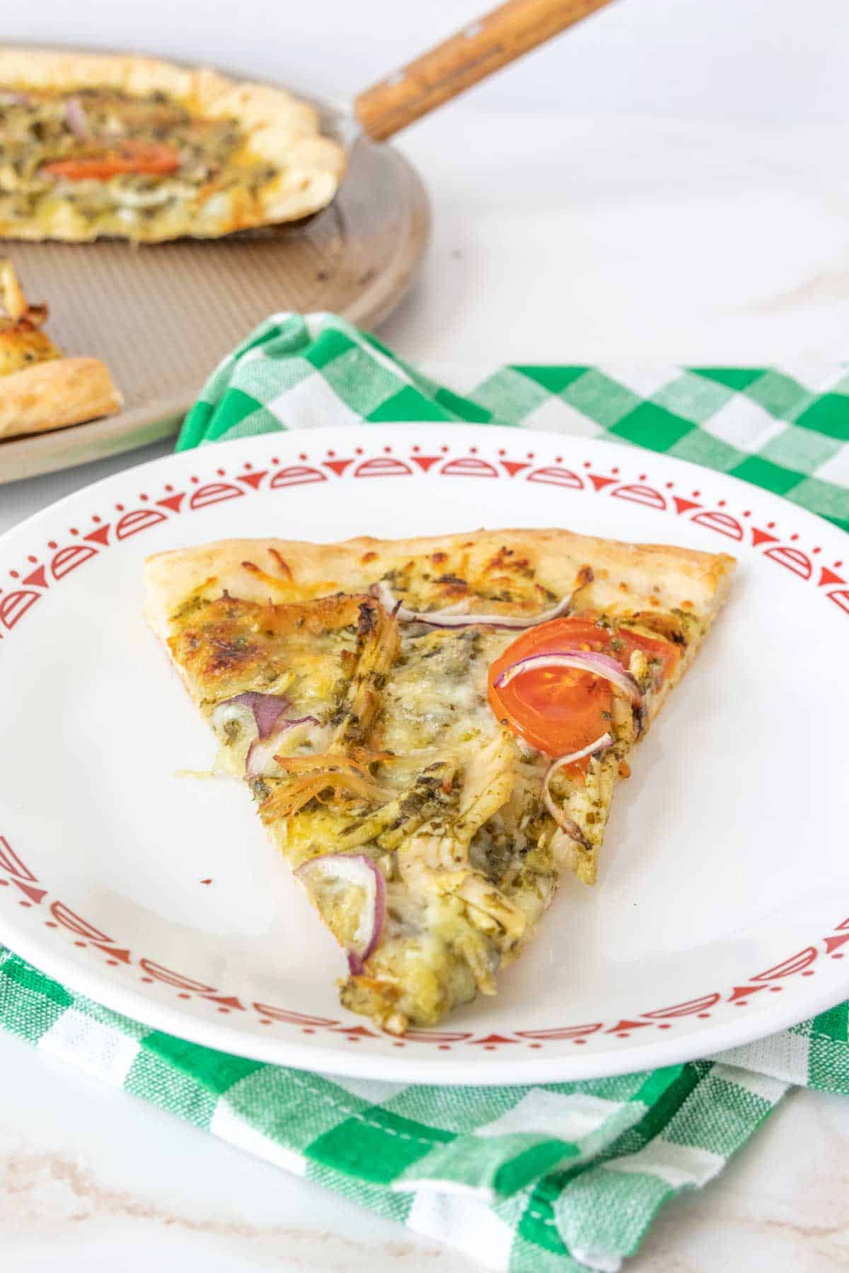 Slice of chicken pesto pizza on a plate with a green plaid napkin underneath.