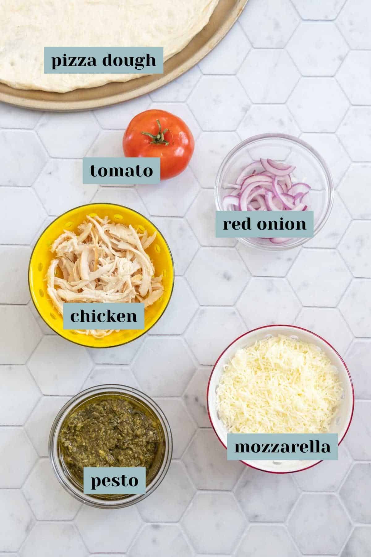 Ingredients for chicken pesto pizza on a tile surface with labels.