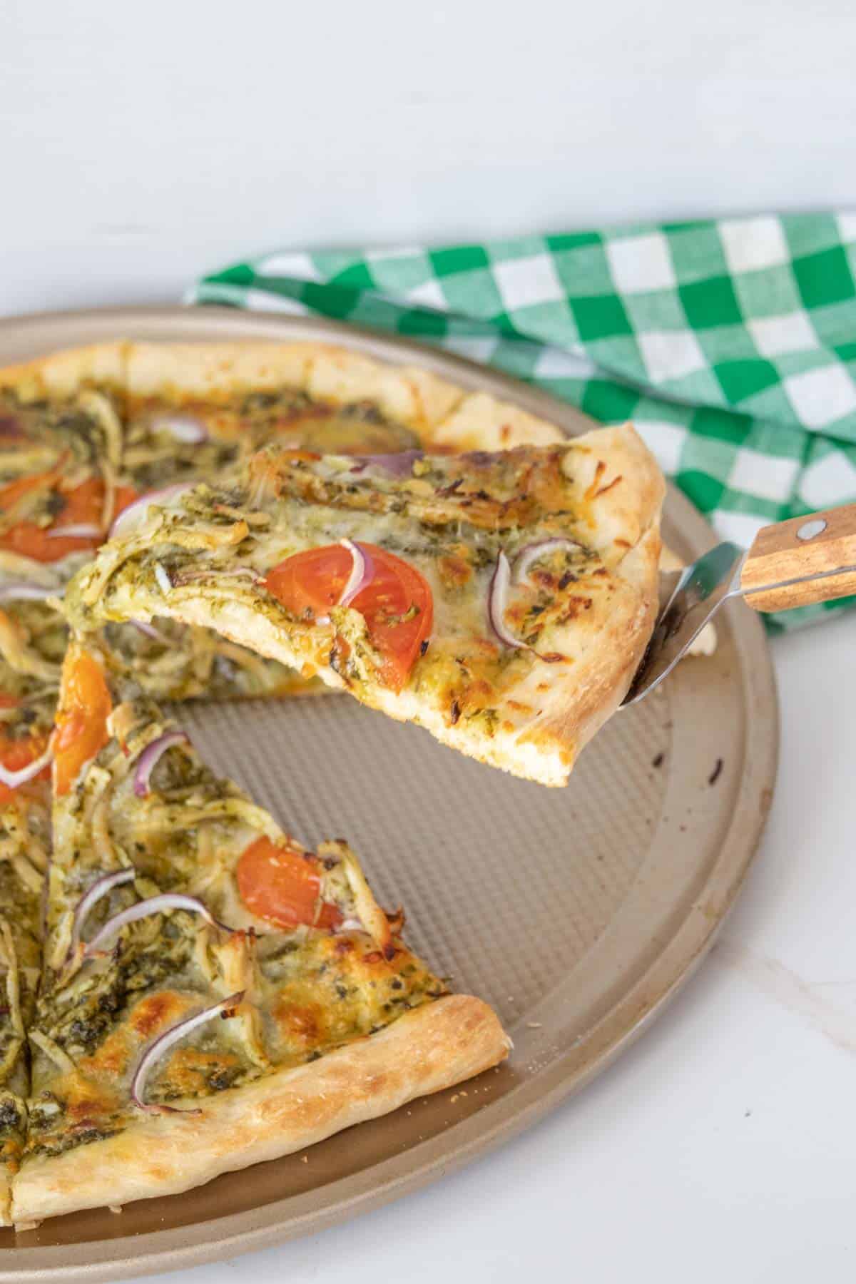 Pizza server holding up slice of chicken pesto pizza from pizza pan.