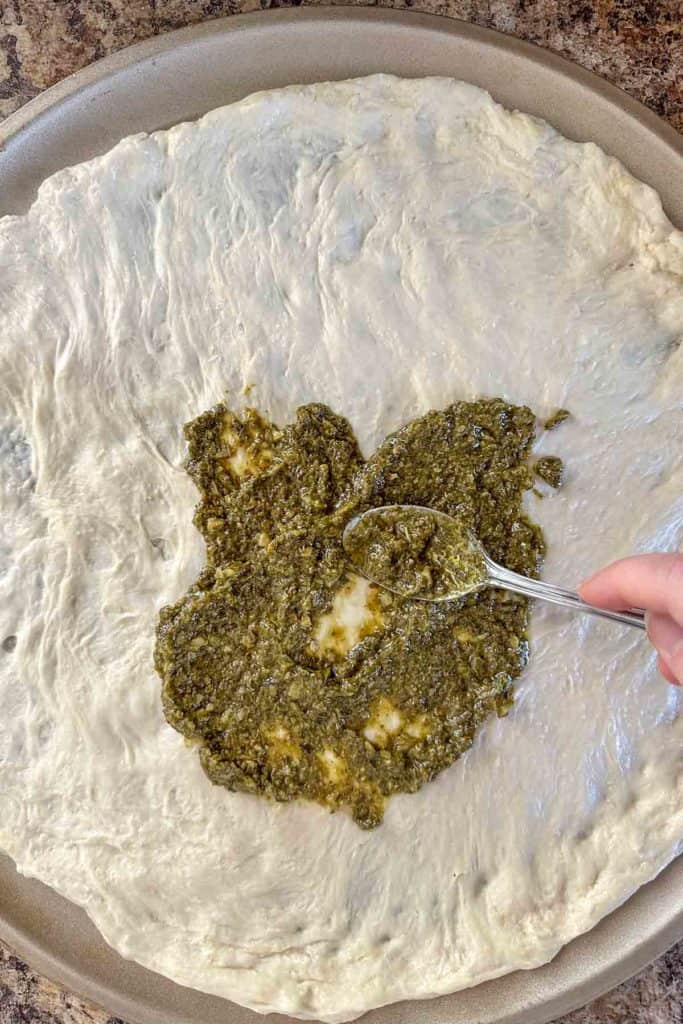 Spreading pesto with a spoon onto unbaked pizza crust.