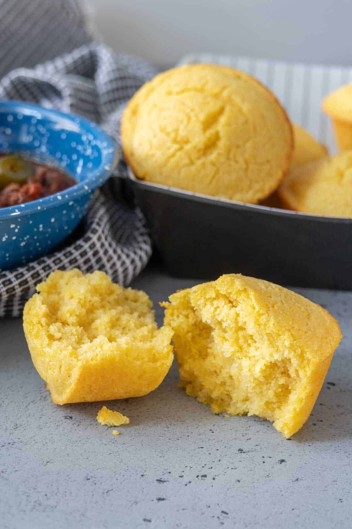 Halved corn muffin on a table with basket of muffins and bowl of chili behind.