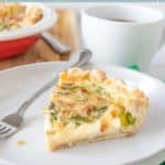 A slice of asparagus quiche on a white plate with a fork, alongside a cup of coffee and another asparagus quiche in the background.