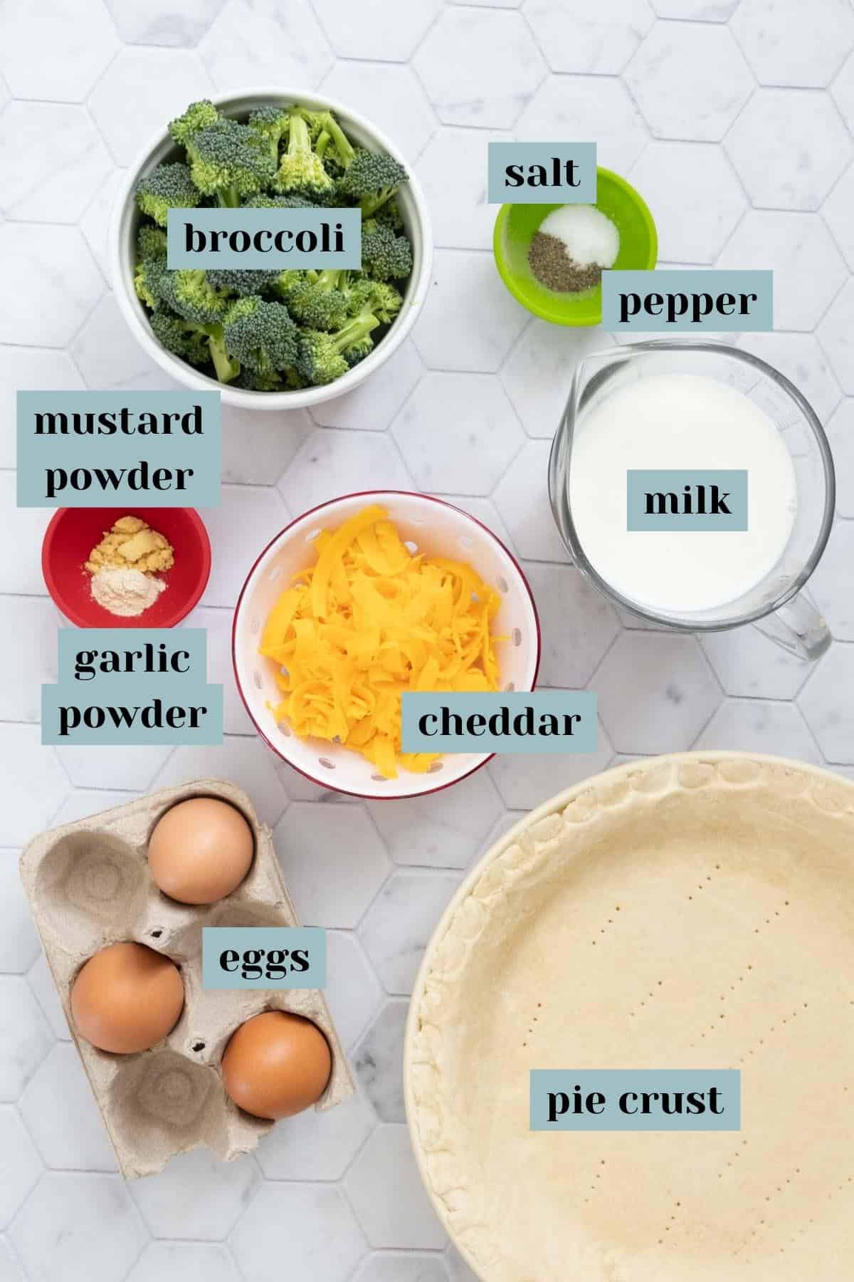 Ingredients for broccoli cheddar quiche on a tile surface with labels.