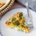 Slice of broccoli cheddar quiche on a white plate with blue rim.