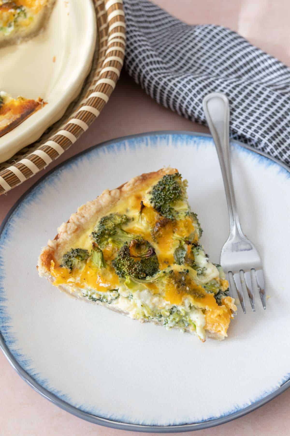 Slice of broccoli cheddar quiche on a white plate with blue rim.