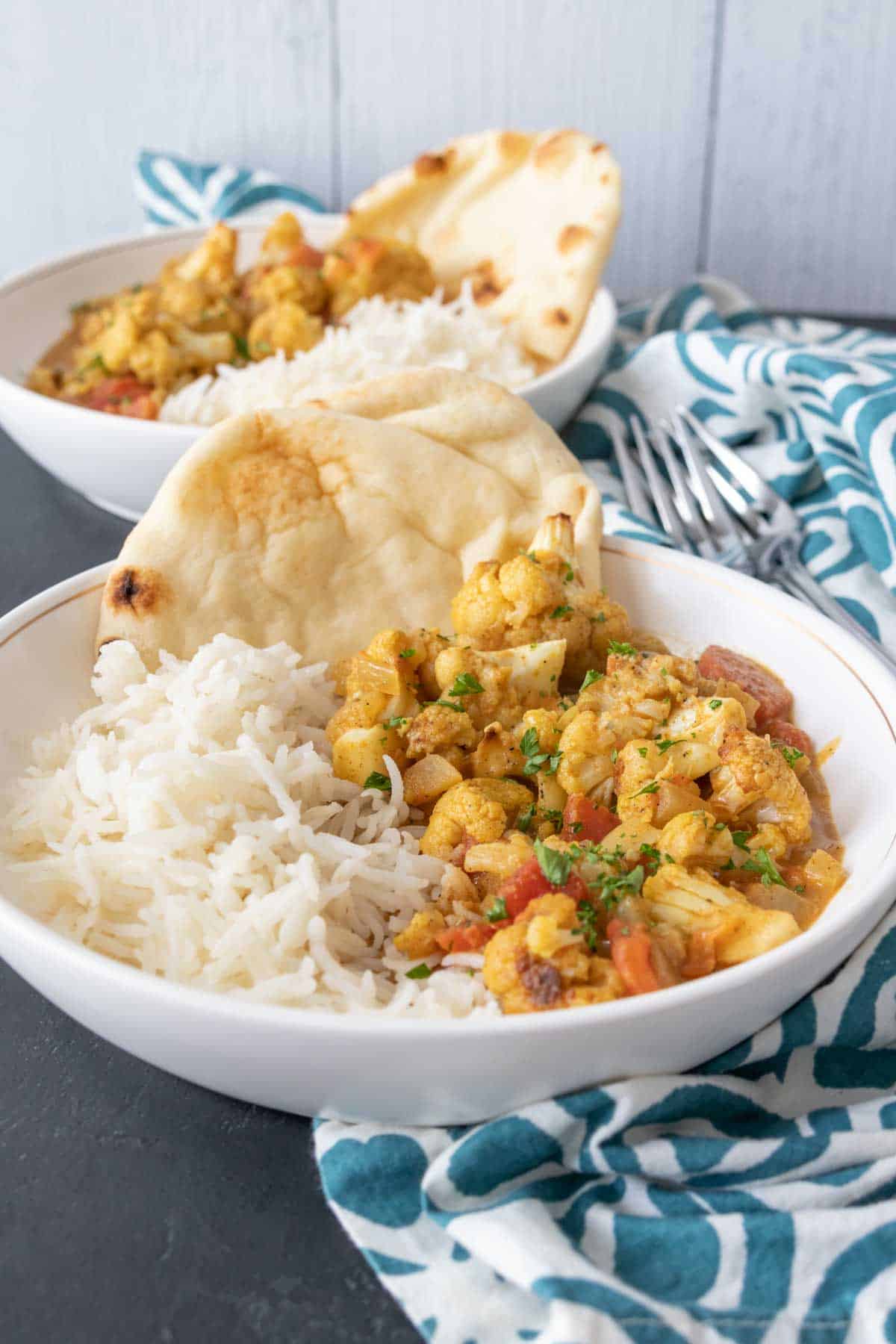 Two white bowls of cauliflower curry, rice, and naan.