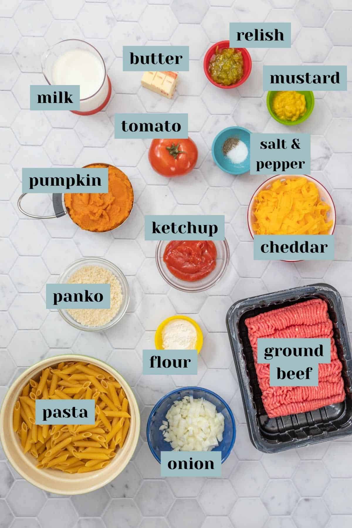 Ingredients for cheeseburger mac and cheese on a tile surface with labels.