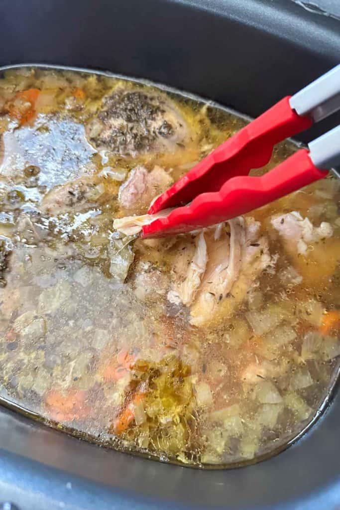 Using tongs to shred cooked chicken thighs in slow cooker.